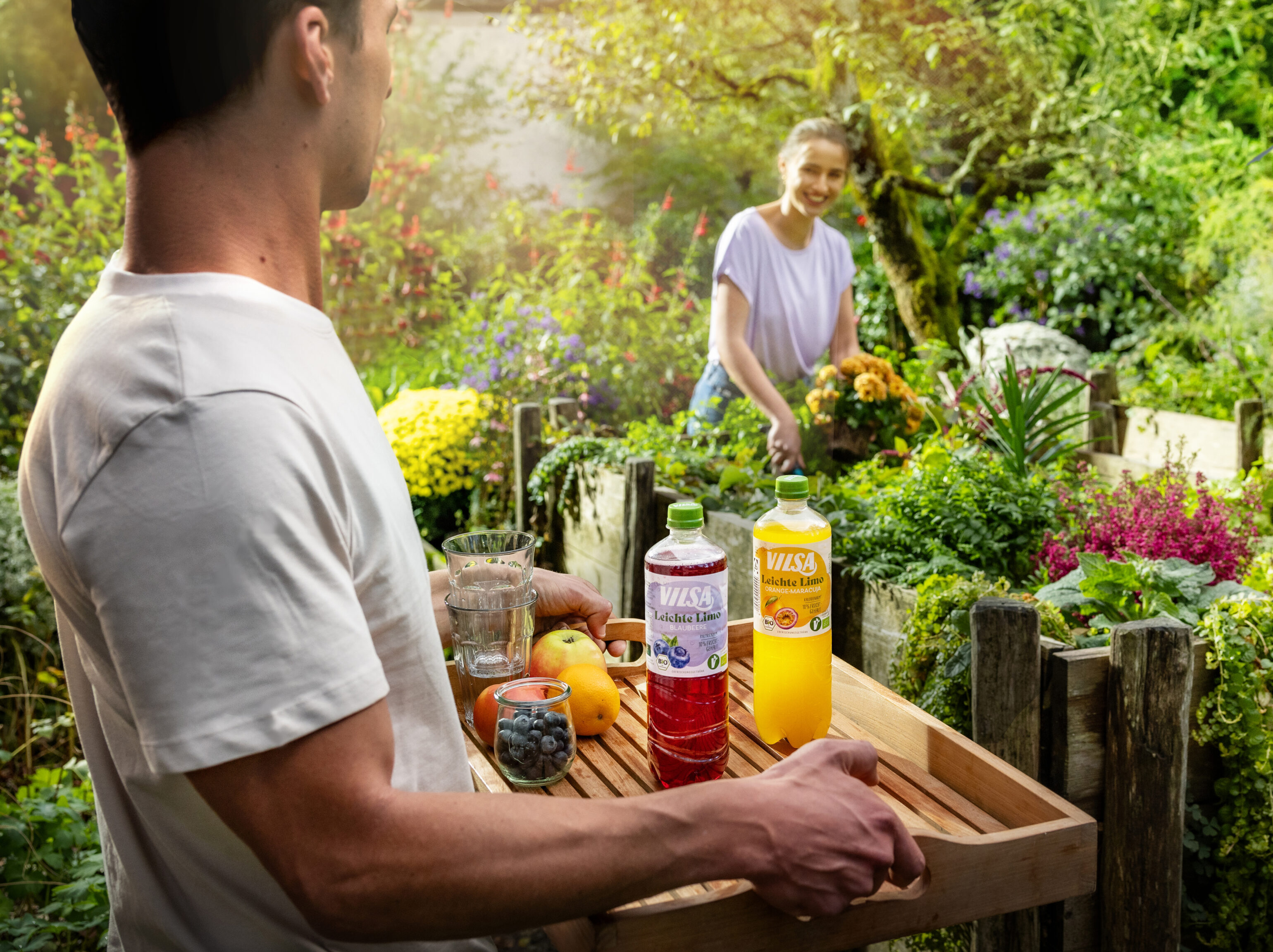 Ein Mann trägt ein Tablett mit Getränken und Obst durch einen farbenfrohen Garten, während eine Frau an einem Hochbeet Blumen pflückt und ihm entgegenlächelt.