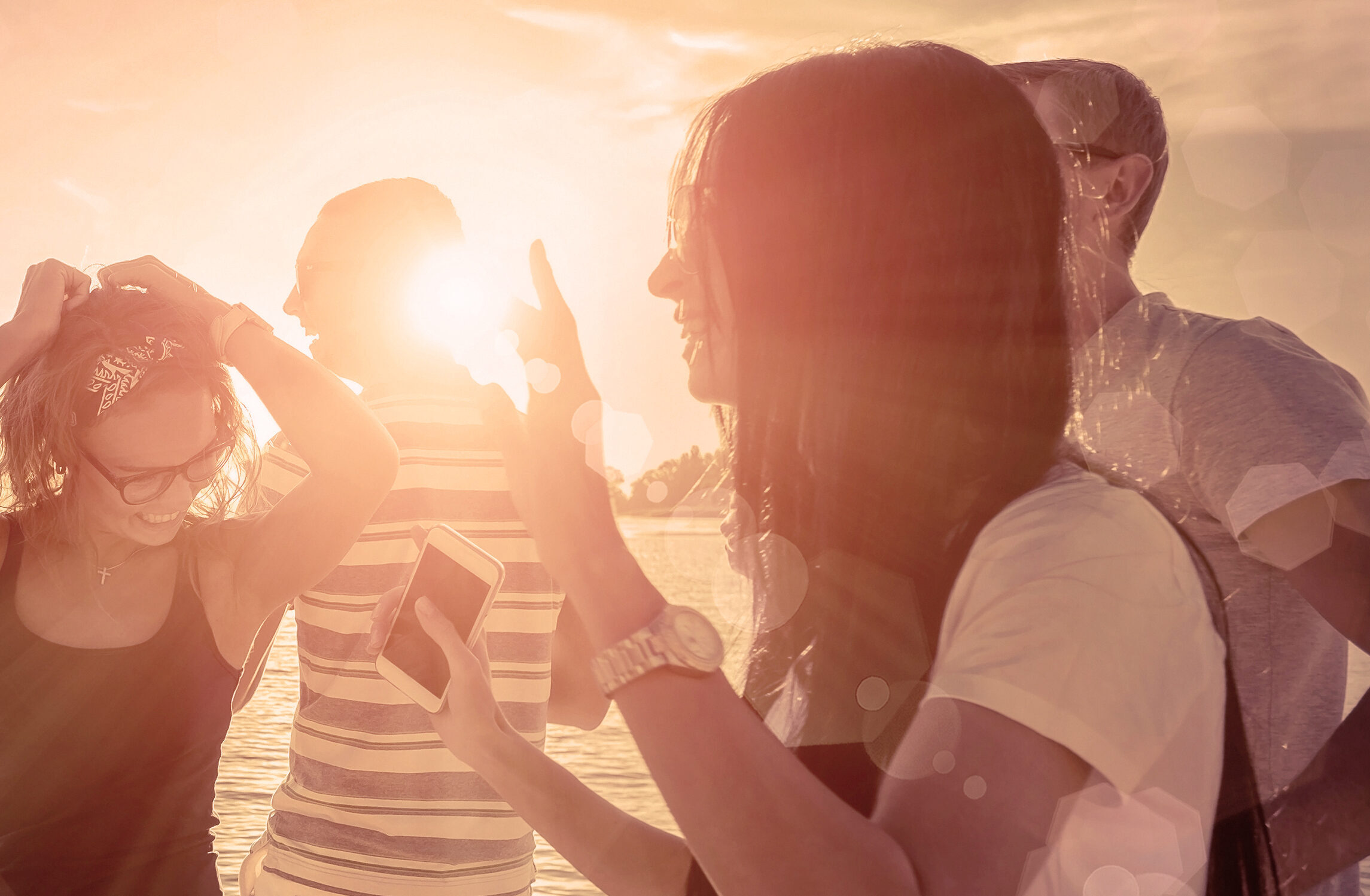 Vier junge Erwachsene genießen gemeinsam den Sonnenuntergang am Wasser. Die Szene ist in warmes, rötliches Sonnenlicht getaucht. Zwei Frauen im Vordergrund lachen und unterhalten sich, eine davon hält ein Smartphone in der Hand. Im Hintergrund stehen zwei Männer, einer trägt ein gestreiftes T-Shirt. Die Atmosphäre wirkt entspannt und sommerlich.