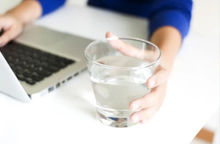 Nahaufnahme von Frauenhänden. Die eine hält ein Glas mit Wasser die andere tippt auf einem Laptop.