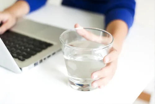 Nahaufnahme von Frauenhänden. Die eine hält ein Glas mit Wasser die andere tippt auf einem Laptop.
