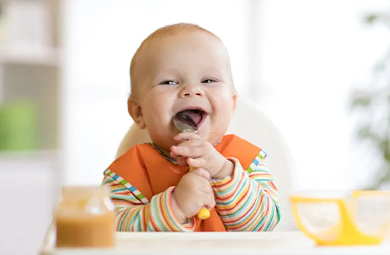 Baby mit Löffel im Mund lächelt in die Kamera.