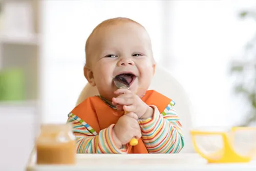 Baby mit Löffel im Mund lächelt in die Kamera.