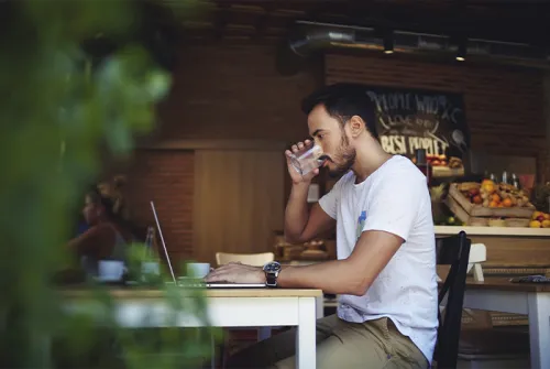 junger Mann sitzt an einem Tisch an einem Laptop und trinkt ein Glas Wasser