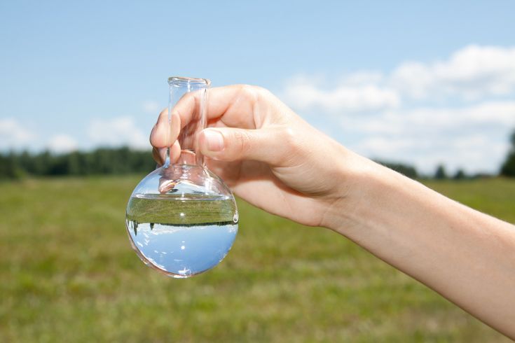 eine Hand hält ein Reagenzglas mit Wasser vor einer grünen Wiese