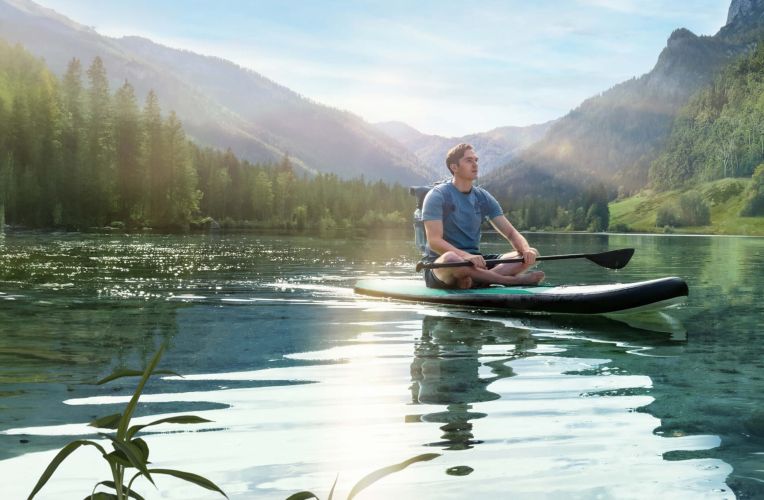 Ein Mann in blauem T-Shirt sitzt im Schneidersitz auf seinem SUP auf einem türkis-blauen See umringt von Bergen und Wäldern. Der Himmel ist blau und die Sonne scheint auf den See.