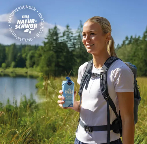 Merle Frohms steht in ihren Sportanziehsachen in der wunderschönen grünen Natur. In der Hand hält sie ein VILSA Tetra Pak Carat Naturelle. Sie lächelt in die Ferne. Im Hintergrund sieht man einen Wald und einen See.