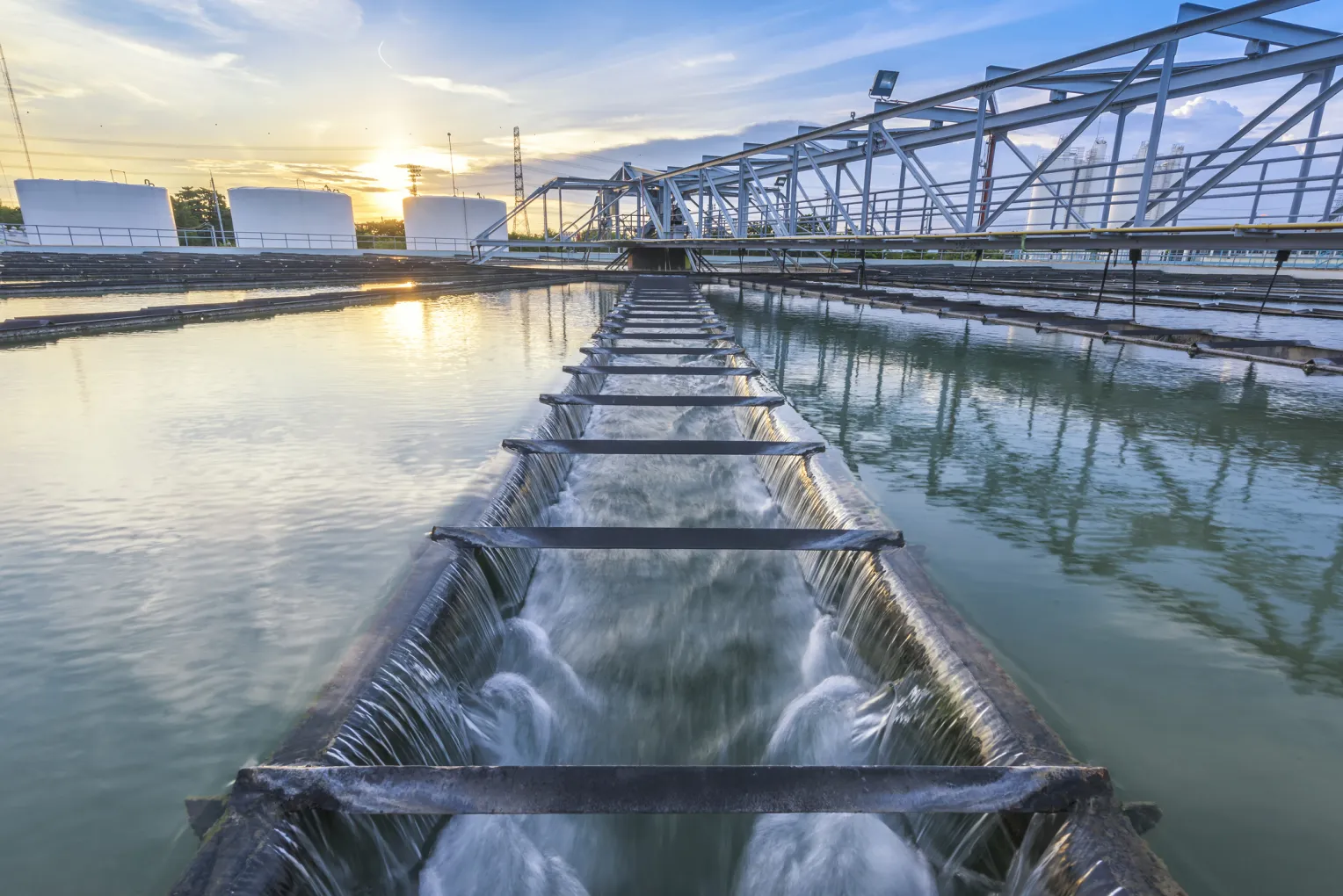 Wasseraufbereitungsanlage bei Sonnenuntergang mit fließendem Wasser durch ein Klärbecken, umgeben von großen weißen Speichertanks und Metallstrukturen.