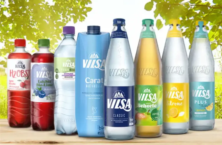 Mehrere VILSA-Getränkeflaschen und -packungen in einer Reihe auf einem Holztisch. Im Hintergrund sonniger, grüner Blätterhimmel. Sorten: VILSA H2Obst, leichte Bio-Limo, PLUS Bio, Carat Naturelle, Classic Mineralwasser, Apfel-Schorle, Zitrone, und Lemon Calcium. Die Verpackungen sind bunt gestaltet in Rot, Lila, Blau, Grün und Gelb.