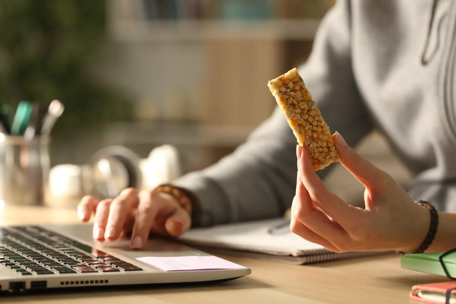 Eine Person arbeitet an einem Laptop und hält in der freien Hand einen Müsliriegel. Auf dem Schreibtisch liegen Notizbücher, Stifte und Kopfhörer.