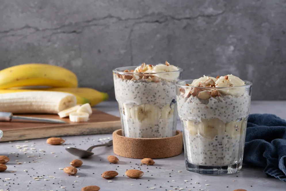 Zwei Gläser mit Chia-Pudding, garniert mit Bananenscheiben und gehackten Mandeln, stehen auf einem grauen Tisch. Im Hintergrund liegen ganze Bananen und ein Schneidebrett mit geschnittenen Bananenscheiben. Mandeln und Chiasamen sind auf dem Tisch verstreut.