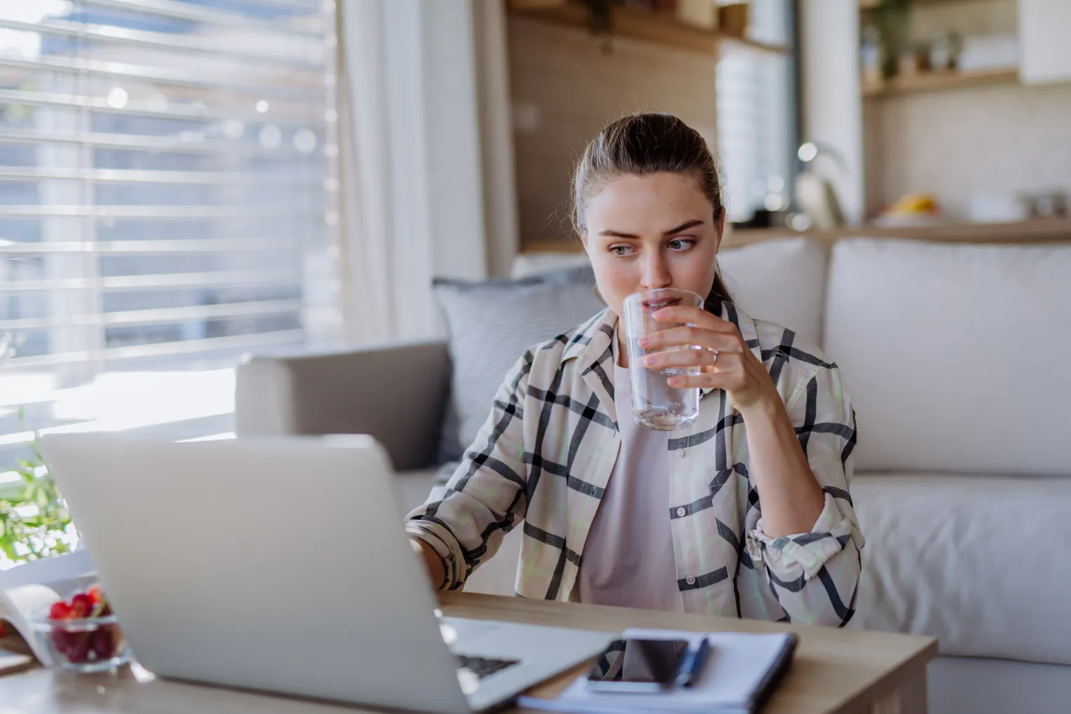 Das Bild zeigt eine junge Frau, die an einem Tisch sitzt und aus einem Glas Wasser trinkt. Vor ihr steht ein aufgeklappter Laptop, daneben liegen ein Notizbuch, ein Smartphone und ein Glas mit Kirschen. Die Frau trägt ein kariertes Hemd und wirkt konzentriert, während sie eine kleine Pause vom Arbeiten oder Lernen macht. Der Hintergrund zeigt eine gemütliche, helle Wohnung mit großen Fenstern, durch die viel Licht hereinfällt.