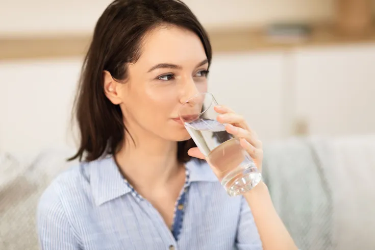 Das Bild zeigt eine junge Frau mit dunklem, schulterlangem Haar, die entspannt ein Glas Wasser trinkt. Sie trägt ein hellblau-weiß gestreiftes Hemd. Der Hintergrund ist weich und unscharf, wodurch der Fokus klar auf der Frau liegt. Die Atmosphäre wirkt ruhig, gemütlich und gesundheitsbewusst.