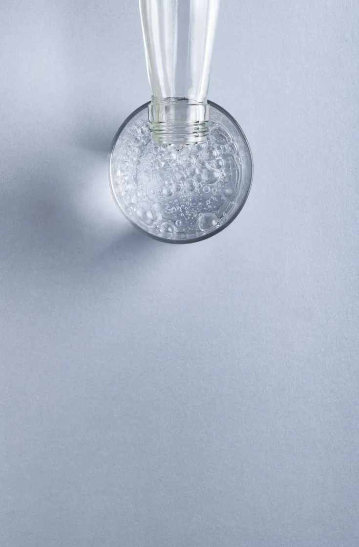 Das Bild zeigt eine Glasflasche, die auf den Kopf gestellt wird und sprudelndes Wasser in ein Glas einschenkt. Die Aufnahme erfolgt von oben. Man sieht viele kleine Luftblasen im Wasser. Der Hintergrund ist einfarbig hellblau und wirkt sehr sauber und minimalistisch. Die Szene vermittelt Frische und Klarheit.