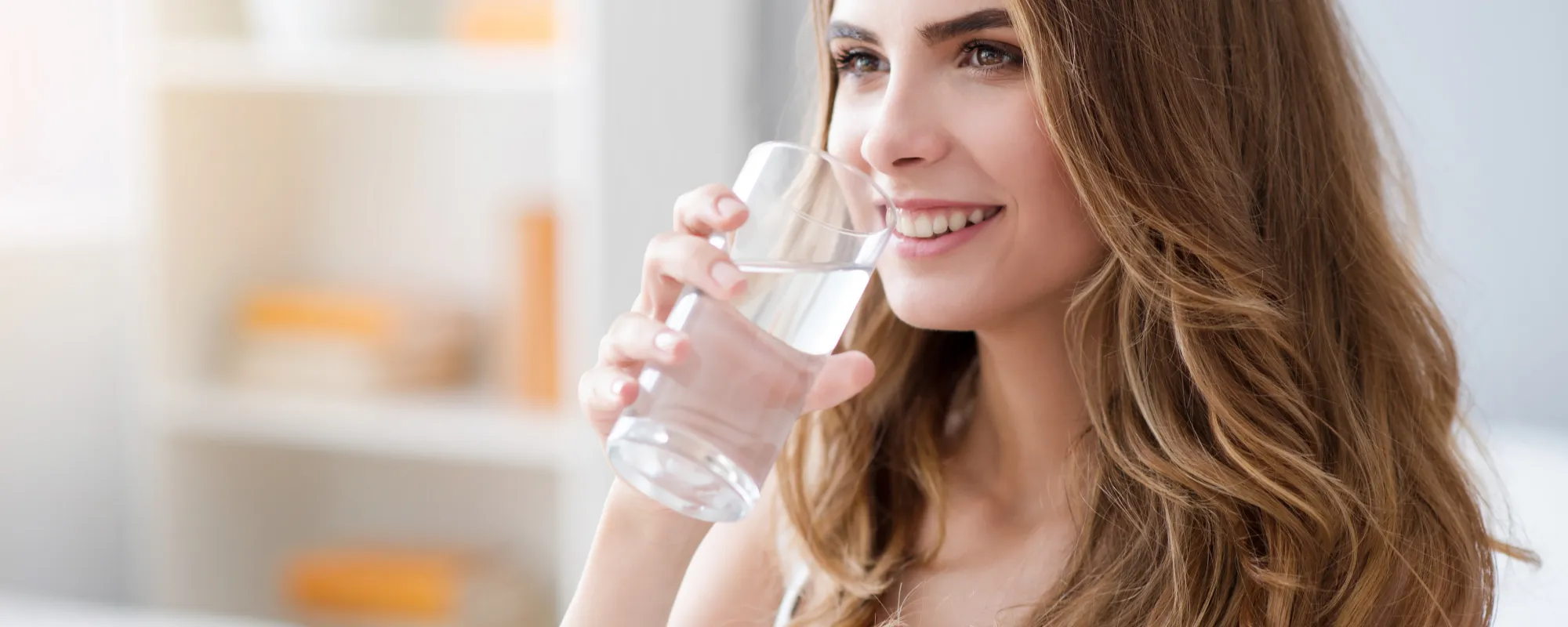 Das Bild zeigt eine junge Frau mit langen, gewellten, braunen Haaren, die lächelnd ein Glas Wasser trinkt. Sie befindet sich in einem hellen, freundlich eingerichteten Raum. Im Hintergrund sieht man ein weißes Regal mit einigen Büchern und einer kleinen Topfpflanze darauf. Die Stimmung des Bildes wirkt frisch, entspannt und gesund.