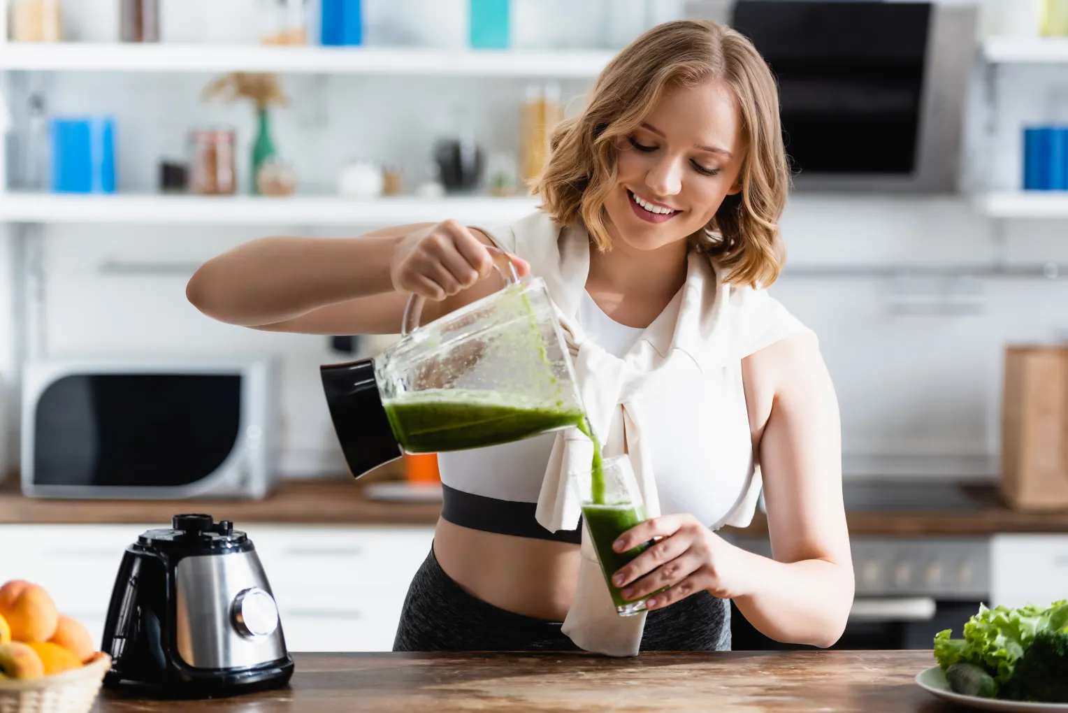 Junge Frau in Sportkleidung steht in einer modernen Küche und gießt einen grünen Smoothie aus einem Mixer in ein Glas. Sie lächelt und wirkt zufrieden.