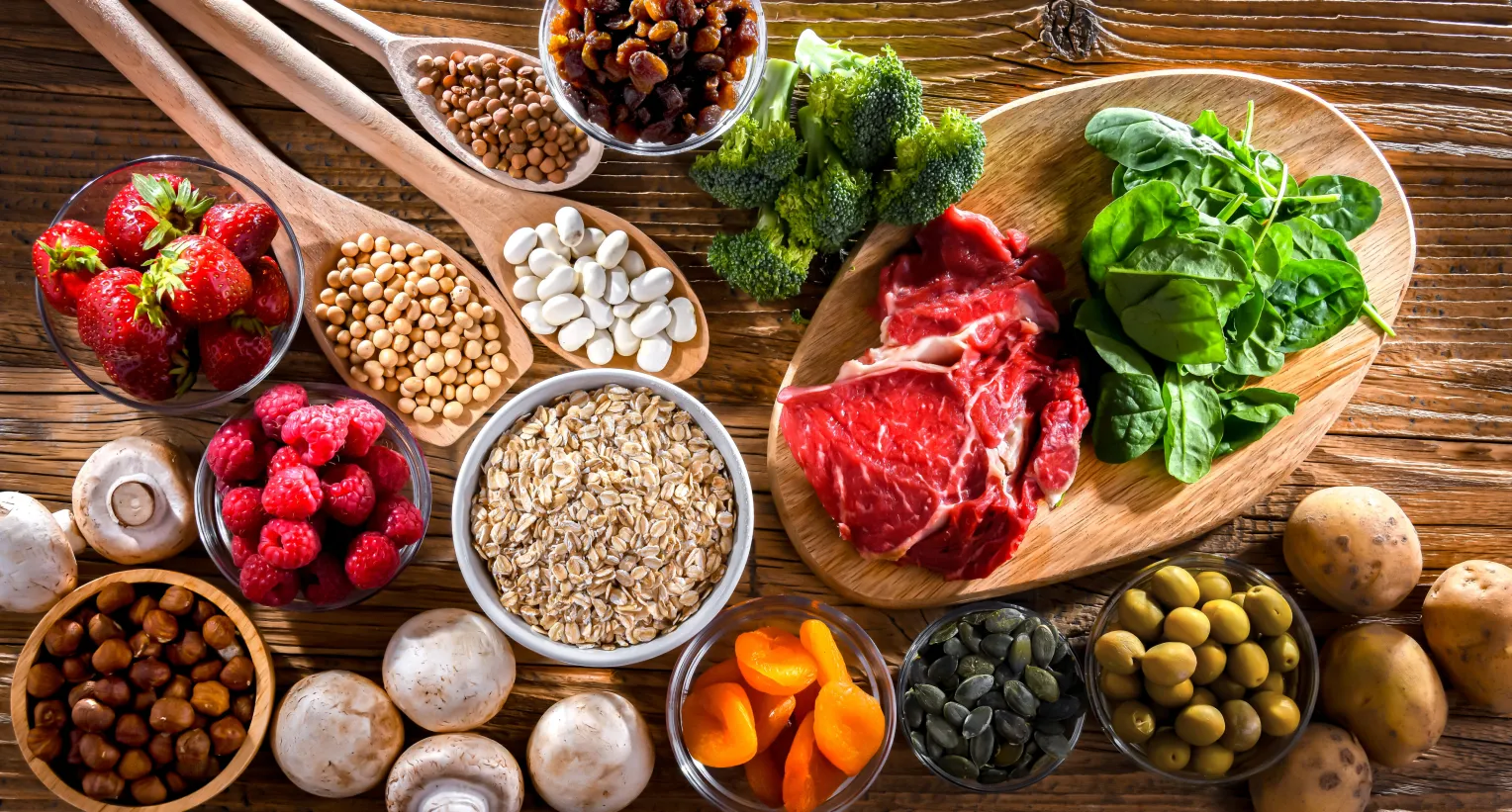 Verschiedene gesunde Lebensmittel auf einem Holztisch: frische Erdbeeren, Himbeeren, Brokkoli, Spinat, rohes rotes Fleisch, weiße Bohnen, Linsen, Sojabohnen, Haferflocken, Haselnüsse, Champignons, Trockenfrüchte, Kürbiskerne, grüne Oliven und Kartoffeln.