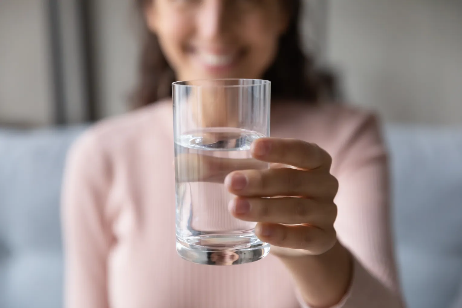Eine Person in einem hellrosa, langärmligen Oberteil lächelt und hält ein Glas Wasser in die Kamera. Der Hintergrund ist unscharf, sodass der Fokus auf dem Glas und der Hand liegt.