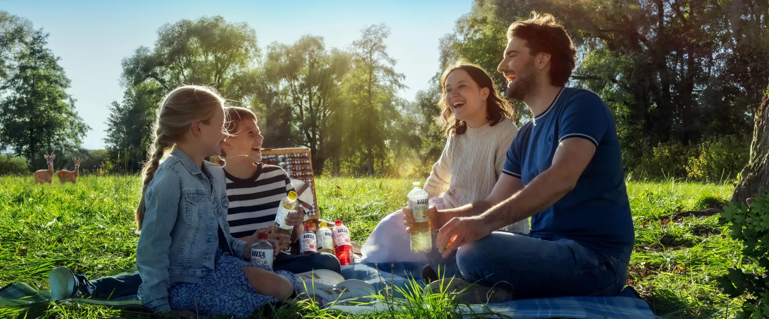 Familie sitzt lachend auf einer Wiese bei einem Picknick, hält Getränkeflaschen in der Hand. Im Hintergrund stehen zwei Rehe am Waldrand. Die Szene spielt an einem sonnigen Tag in der Natur.