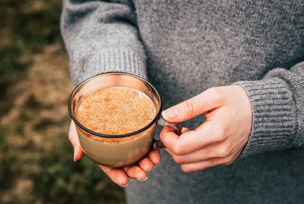 Person in grauem Wollpullover hält eine Tasse mit einem warmen Getränk, bestreut mit Zimt oder Kakaopulver, in beiden Händen. Der Hintergrund ist unscharf und zeigt eine natürliche Umgebung.