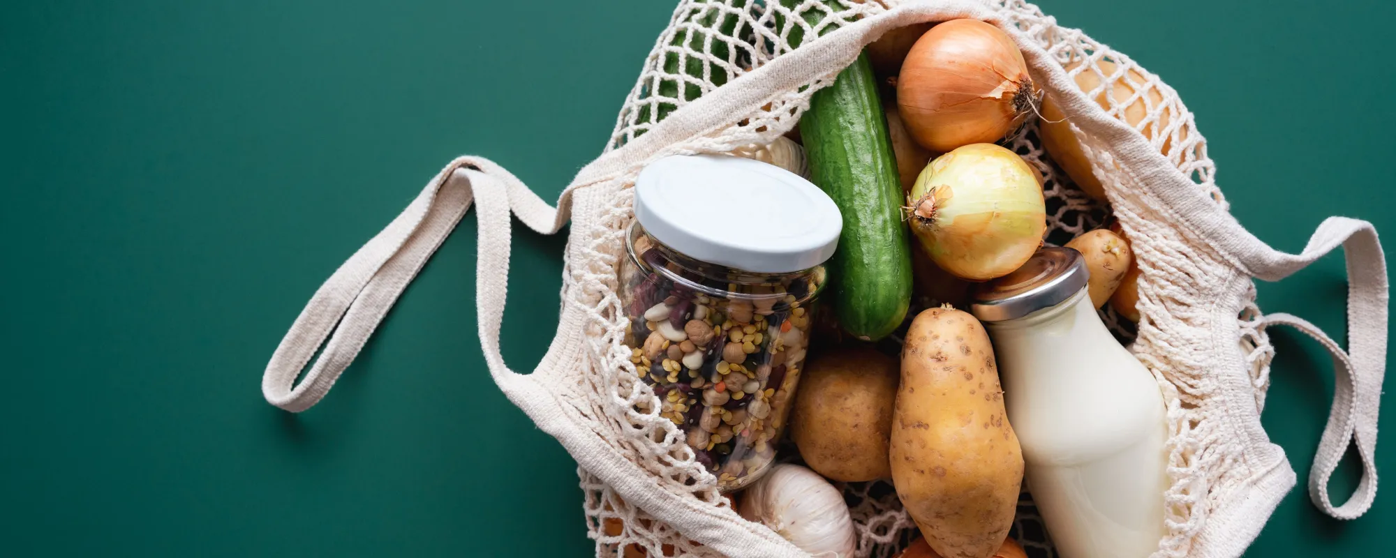 Wiederverwendbare Einkaufstasche aus Baumwolle, gefüllt mit frischen Lebensmitteln: Kartoffeln, Zwiebeln, Gurke, Knoblauch, einer Glasflasche Milch und einem Glas mit getrockneten Hülsenfrüchten, auf grünem Hintergrund.