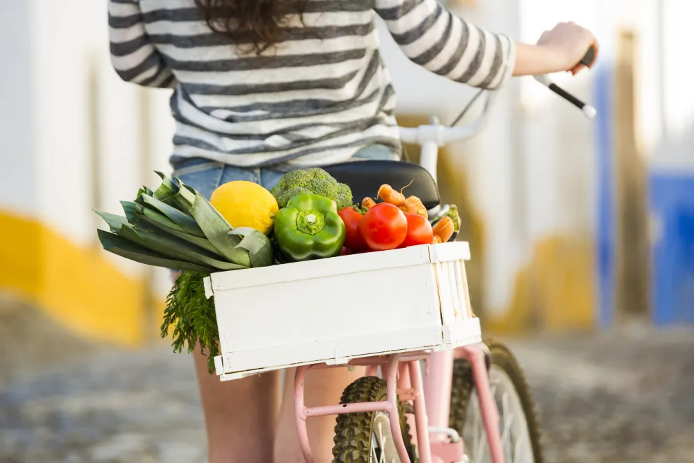 Person fährt Fahrrad mit einer Holzkiste voller frischem Gemüse wie Lauch, Paprika, Tomaten, Brokkoli und Karotten auf dem Gepäckträger, auf einer sonnigen Kopfsteinpflasterstraße.
