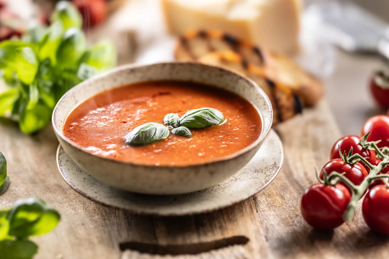 Eine schön angerichtete vegetarische Tomatensuppe mit Parmesan und Basilikum garniert. Neben der Schale liegen Tomaten, Basilikum und Brot.