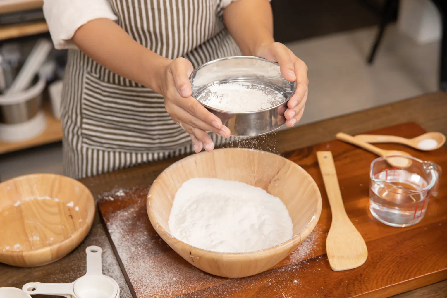 Ansicht von Frauenhänden, die Mehl in eine Holzschüssel sieben, bevor der Teig geknetet wird. Neben der Schüssel liegen Holzlöffel, ein Glas mit Wasser und weiteres Backzubehör.