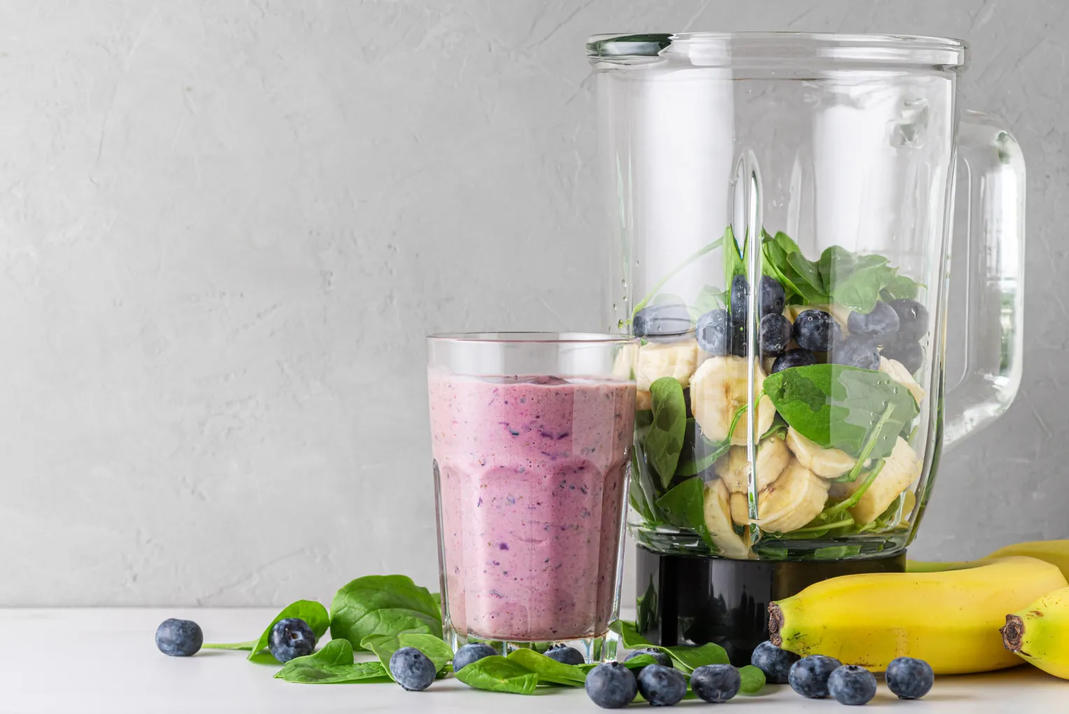 Glas mit Blaubeeren-, Bananen- und Spinatsmoothie. Daneben steht ein Mixer mit frischen, saftigen Zutaten.