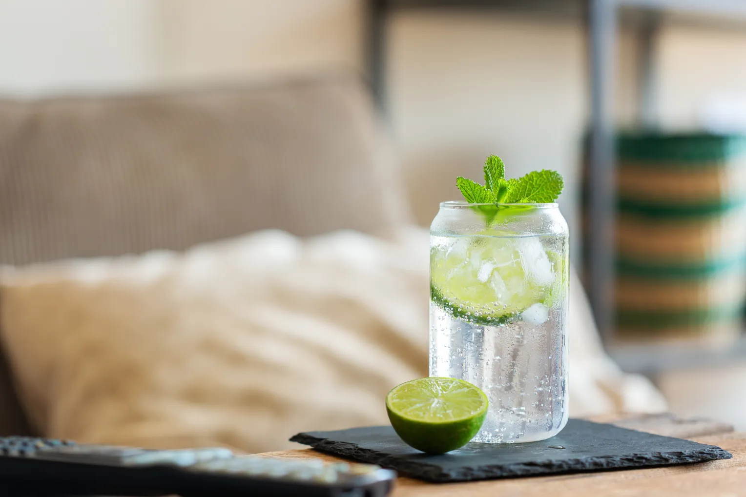 Ein Glas mit frischen Mineralwasser, Eiswürfeln, Limettenscheiben und Minze auf einem Holztisch. Im Hintergrund sieht man ein gemütliches Wohnzimmer.