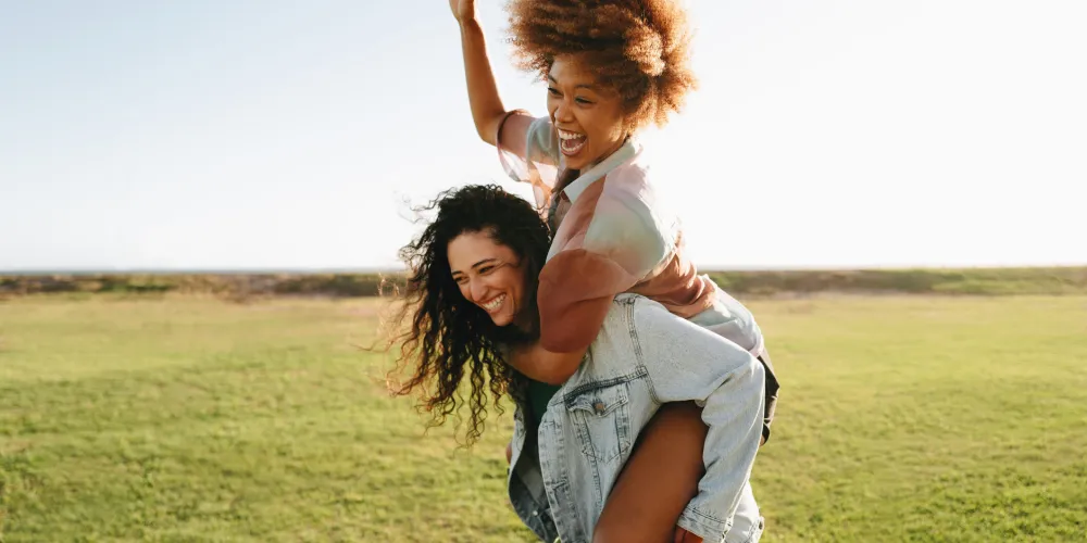 Zwei junge Frauen haben Spaß im Freien. Eine trägt die andere auf dem Rücken, beide lachen ausgelassen. Sie befinden sich auf einer grünen Wiese unter klarem, blauem Himmel. Die Szene strahlt Freude, Leichtigkeit und Freundschaft aus.