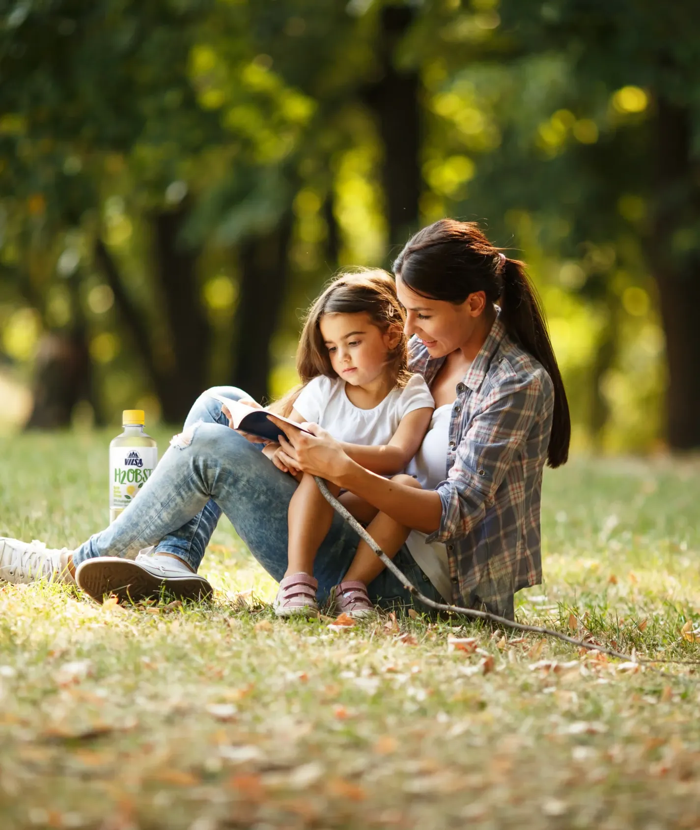 Eine Frau sitzt mit einem kleinen Mädchen auf einer Wiese im Park und liest ihr aus einem Buch vor. Im Hintergrund sind Bäume und eine Flasche Wasser zu sehen. Die Szene wirkt ruhig und harmonisch.