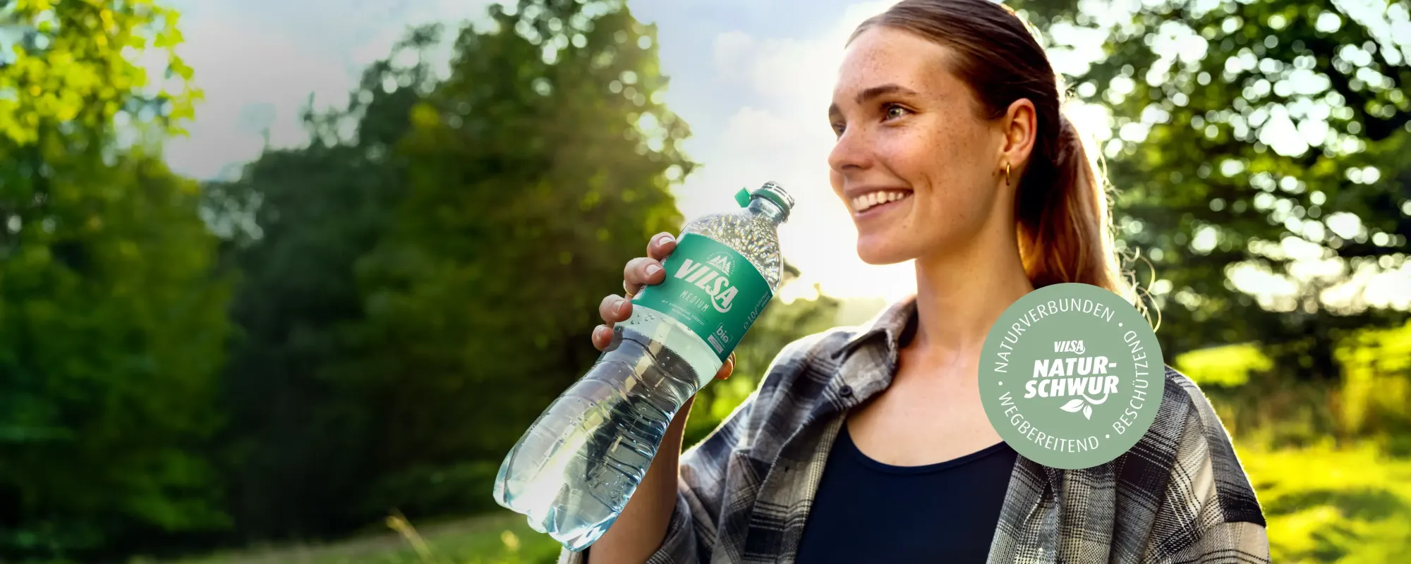 Junge Frau steht in der Natur und lächelt, während sie eine Flasche Vilsa Mineralwasser in der Hand hält. Im Hintergrund sind Bäume und Sonnenlicht zu sehen, vorne rechts ein Logo mit der Aufschrift ‚Naturschwur‘.