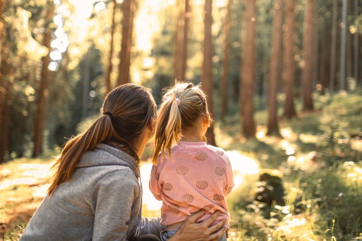 Eine Frau und ein kleines Mädchen mit blonden Zöpfen sitzen nebeneinander im Wald und schauen gemeinsam in die Ferne. Die Szene ist in warmes Sonnenlicht getaucht, das durch die hohen Bäume fällt. Die Frau trägt einen grauen Hoodie, das Mädchen ein rosa Oberteil mit Muster. Die Stimmung wirkt ruhig, innig und naturverbunden.