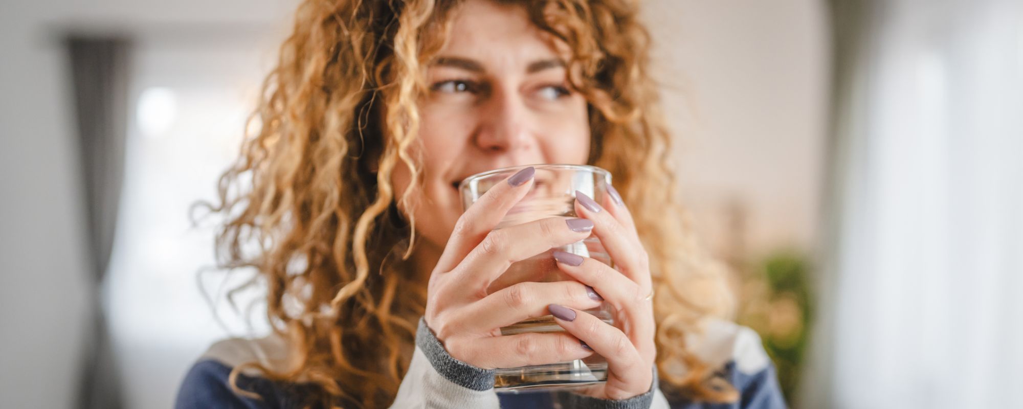 Junge Frau mit lockigem Haar hält ein Glas Wasser mit beiden Händen und lächelt. Sie trägt ein gestreiftes Sweatshirt und befindet sich in einem hellen, gemütlichen Raum.