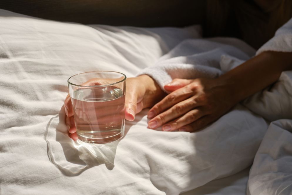 Hand hält ein Glas Wasser auf einem weißen Bettlaken in sanftem Sonnenlicht. Eine zweite Hand ruht entspannt daneben.