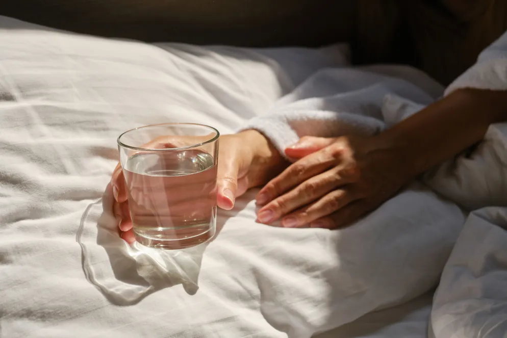 Hand hält ein Glas Wasser auf einem weißen Bettlaken in sanftem Sonnenlicht. Eine zweite Hand ruht entspannt daneben.