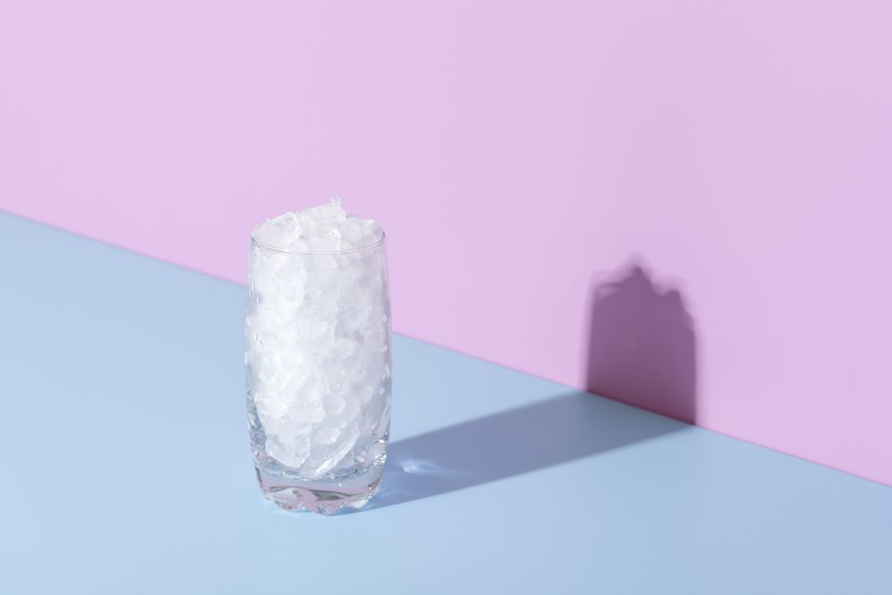 Ein Glas, gefüllt mit Crushed Ice, steht auf einer zweifarbigen Oberfläche mit hellblauer Unterseite und rosa Hintergrund. Das Glas wirft einen deutlichen Schatten auf die Wand, und Kondenswasser ist an der Außenseite sichtbar. Die Szene ist minimalistisch und farblich kontrastreich gestaltet.