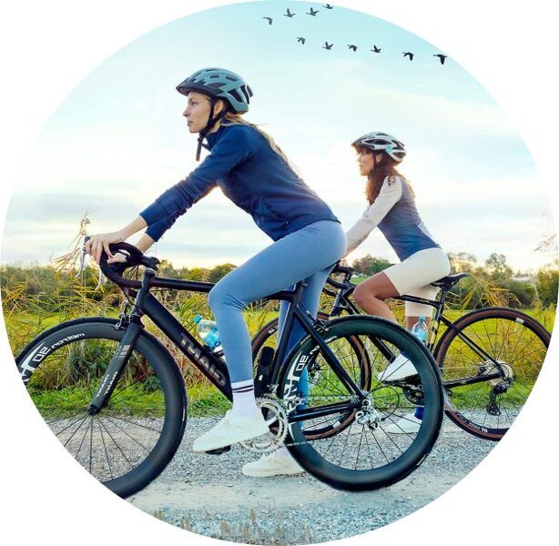 Zwei Frauen fahren bei Sonnenuntergang auf einem Schotterweg Fahrrad. Sie tragen Sportkleidung und Helme. Im Hintergrund fliegen Vögel am Himmel und es ist grüne Natur zu sehen.