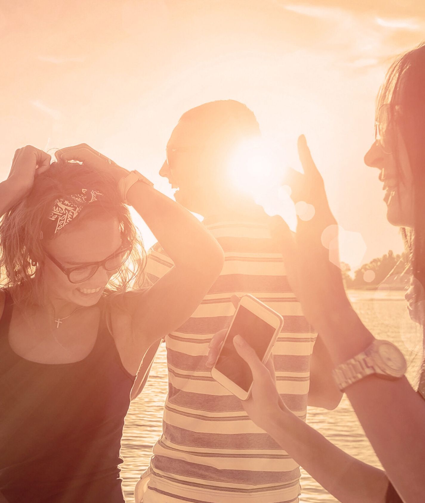 Vier junge Erwachsene genießen gemeinsam den Sonnenuntergang am Wasser. Die Szene ist in warmes, rötliches Sonnenlicht getaucht. Zwei Frauen im Vordergrund lachen und unterhalten sich, eine davon hält ein Smartphone in der Hand. Im Hintergrund stehen zwei Männer, einer trägt ein gestreiftes T-Shirt. Die Atmosphäre wirkt entspannt und sommerlich.