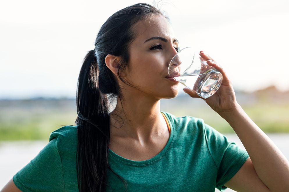 Junge Frau in Sportkleidung trinkt draußen ein Glas Wasser, mit konzentriertem Blick zur Seite.