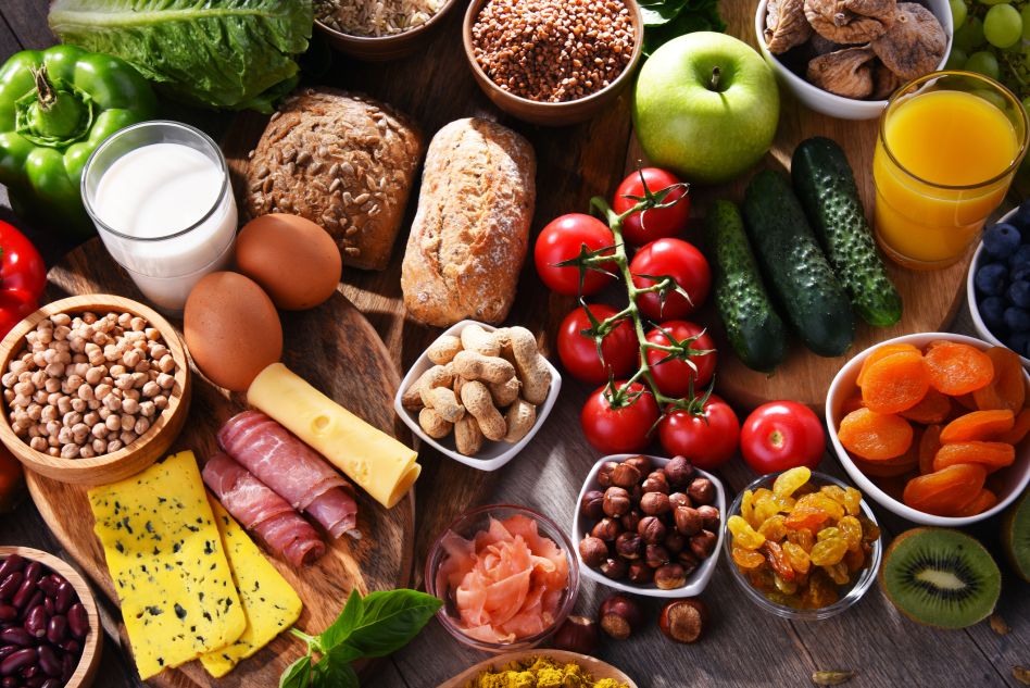 Vielfältige Auswahl gesunder Lebensmittel, darunter frisches Gemüse, Obst, Vollkornbrot, Nüsse, Käse, Hülsenfrüchte, Milch, Eier und Saft, arrangiert auf einem Holztisch.