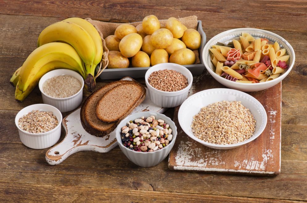 Kohlenhydratreiche Lebensmittel wie Bananen, Kartoffeln, Vollkornbrot, Reis, Hülsenfrüchte, Haferflocken und bunte Nudeln auf einem rustikalen Holztisch.