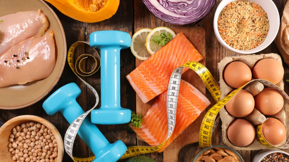 Proteinreiche Lebensmittel wie Lachs, Hühnchen, Eier, Hülsenfrüchte und Nüsse, kombiniert mit Hanteln und Maßband als Symbol für gesunde Ernährung und Fitness.