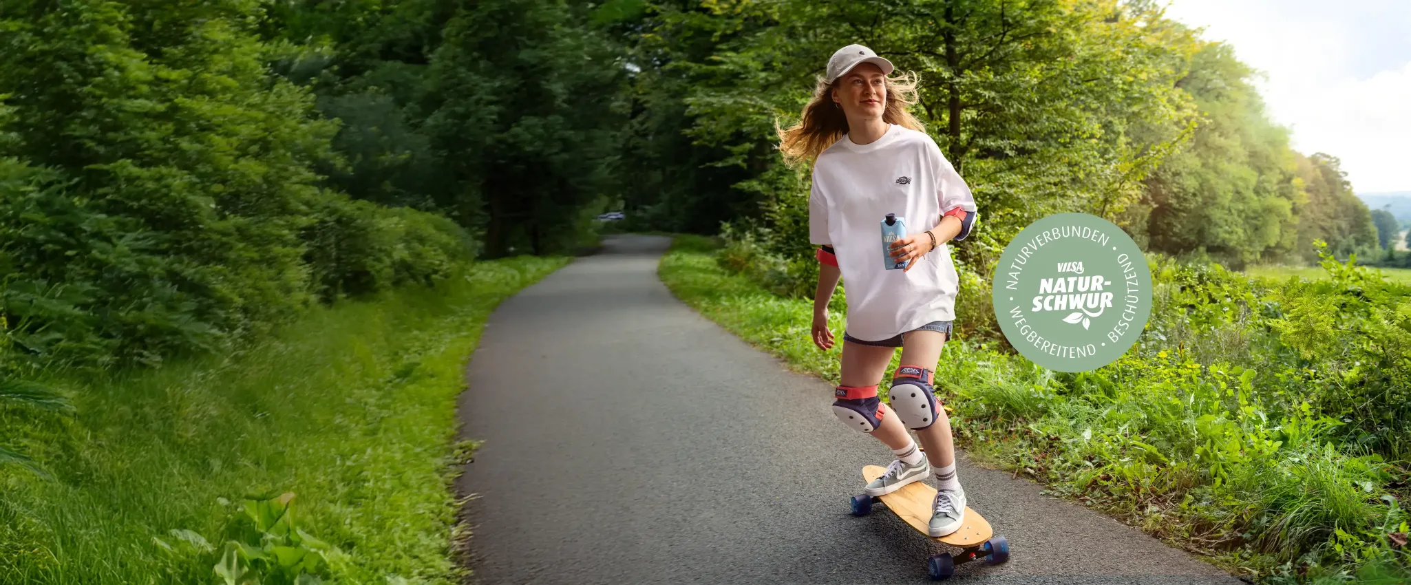 Eine junge Frau fährt bei sonnigem Wetter mit einem Longboard auf einem asphaltierten Weg durch einen grünen Wald. Sie trägt einen weißen Oversize-Pullover, eine Baseballkappe und Schutzausrüstung an Knien und Ellenbogen. In der Hand hält sie einen Getränkekarton von Vilsa. Rechts im Bild ist ein rundes Siegel mit der Aufschrift „VILSA Naturschwur – naturverbunden, wegbereitend, beschützend“ zu sehen.