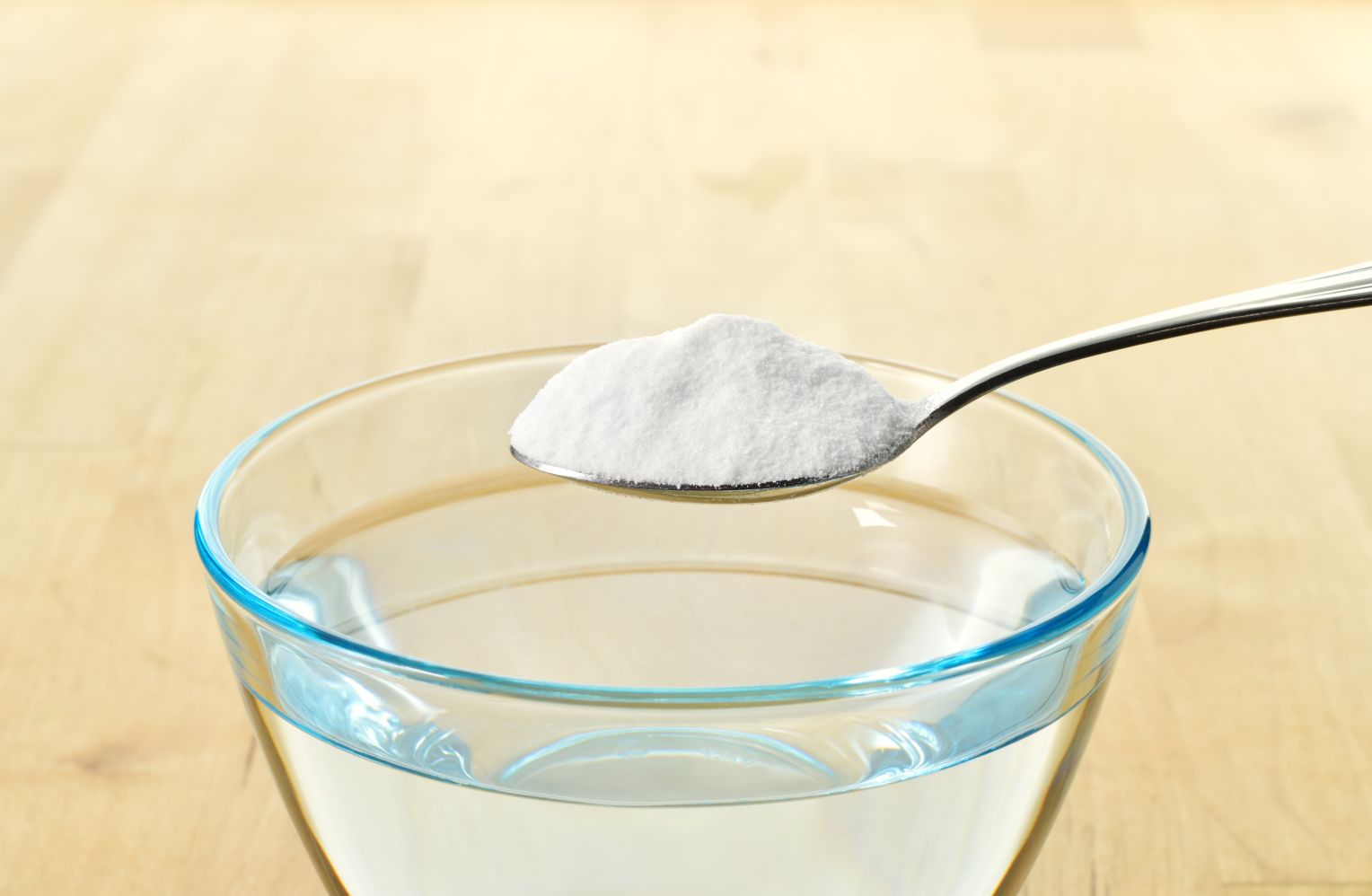 Eine Glasschüssel mit klarem Wasser steht auf einem hellen Holztisch. Über der Schüssel hält ein Löffel eine weiße, pulverartige Substanz – vermutlich Natron oder Backpulver.