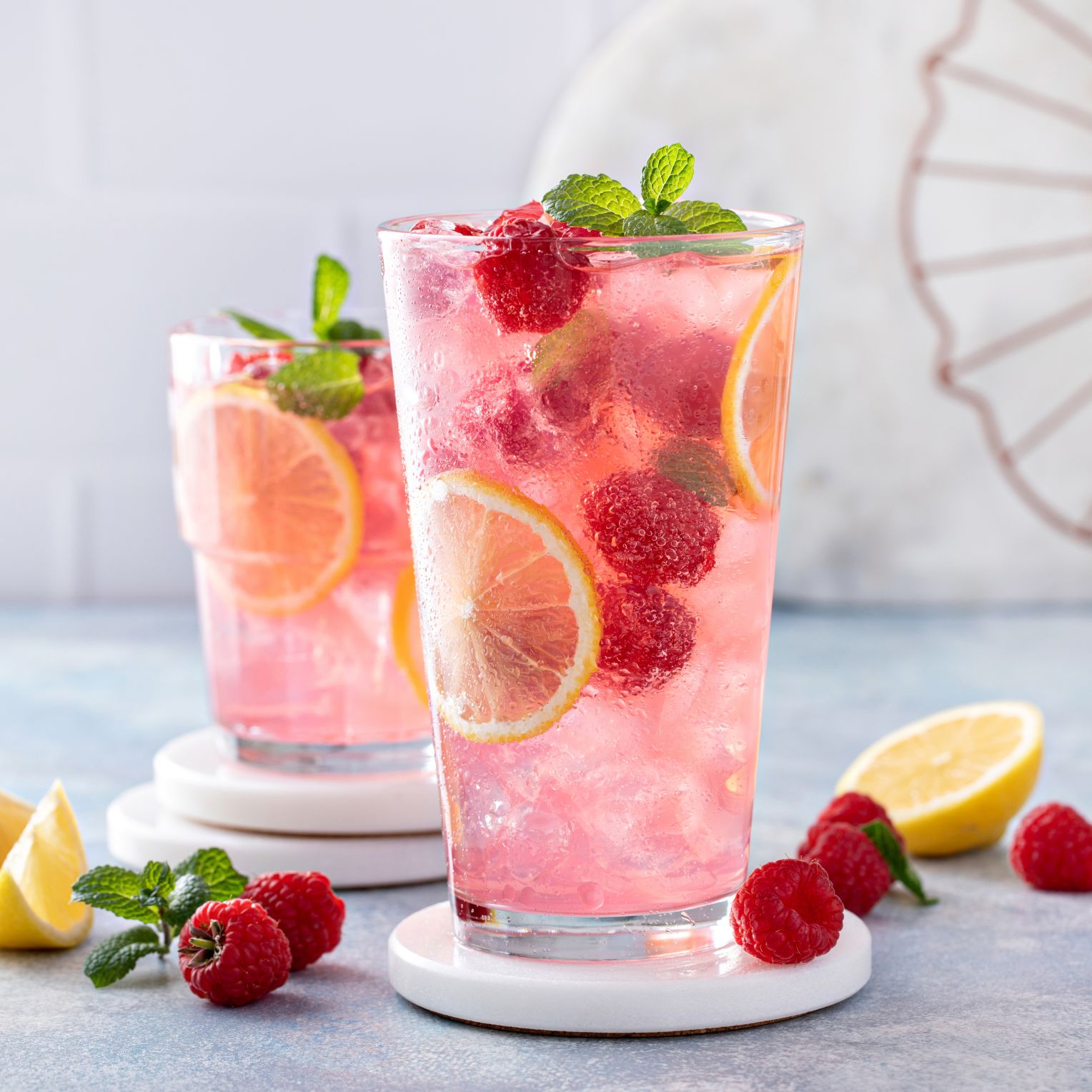 Zwei Gläser mit einem erfrischenden, rosa Getränk stehen auf weißen Untersetzern. Die Getränke enthalten Eiswürfel, Zitronenscheiben, Himbeeren und frische Minzblätter. Im Hintergrund liegen weitere Himbeeren, Minzblätter und eine halbierte Zitrone.