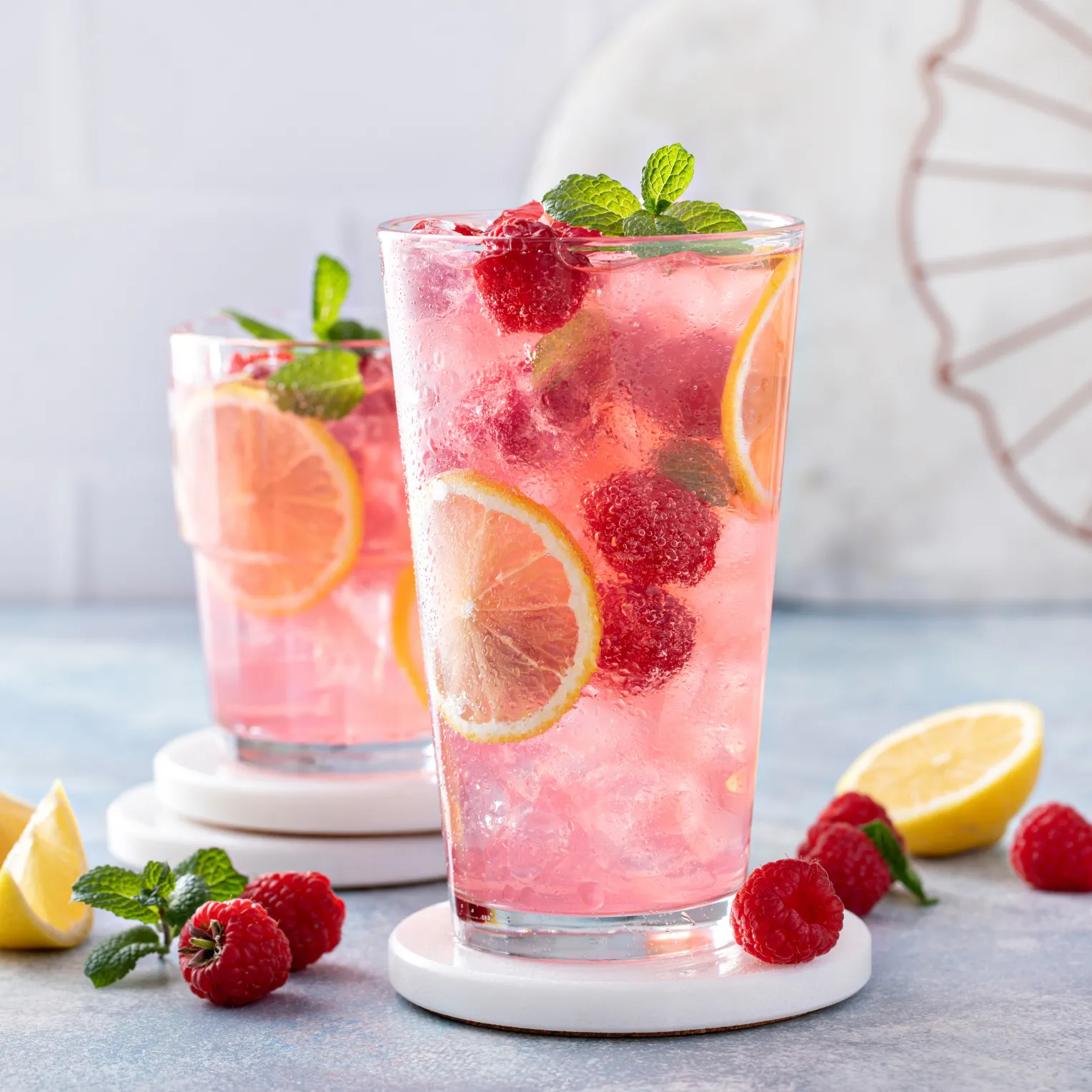 Zwei Gläser mit einem erfrischenden, rosa Getränk stehen auf weißen Untersetzern. Die Getränke enthalten Eiswürfel, Zitronenscheiben, Himbeeren und frische Minzblätter. Im Hintergrund liegen weitere Himbeeren, Minzblätter und eine halbierte Zitrone.