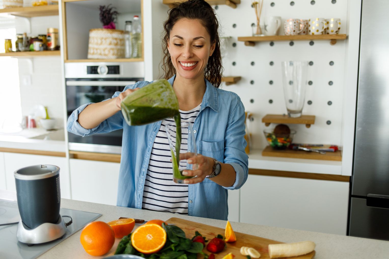 Eine junge Frau steht in einer modernen Küche und gießt einen grünen Smoothie aus einem Mixer in ein Glas. Auf der Arbeitsplatte liegen frische Früchte und Gemüse wie Orangen, Bananen, Erdbeeren und Spinat.