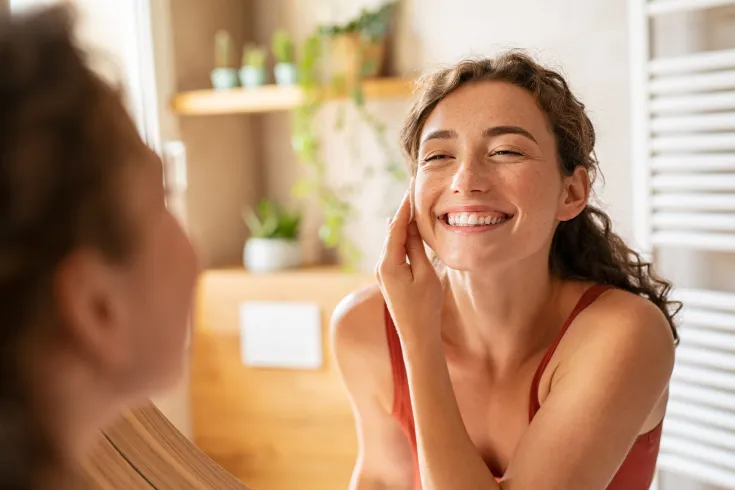Eine lächelnde Frau trägt im Badezimmer eine Creme oder Lotion auf ihr Gesicht auf. Sie steht vor dem Spiegel, wirkt glücklich und pflegt ihre Haut. Im Hintergrund sind Pflanzen und Badezimmerdekoration sichtbar.