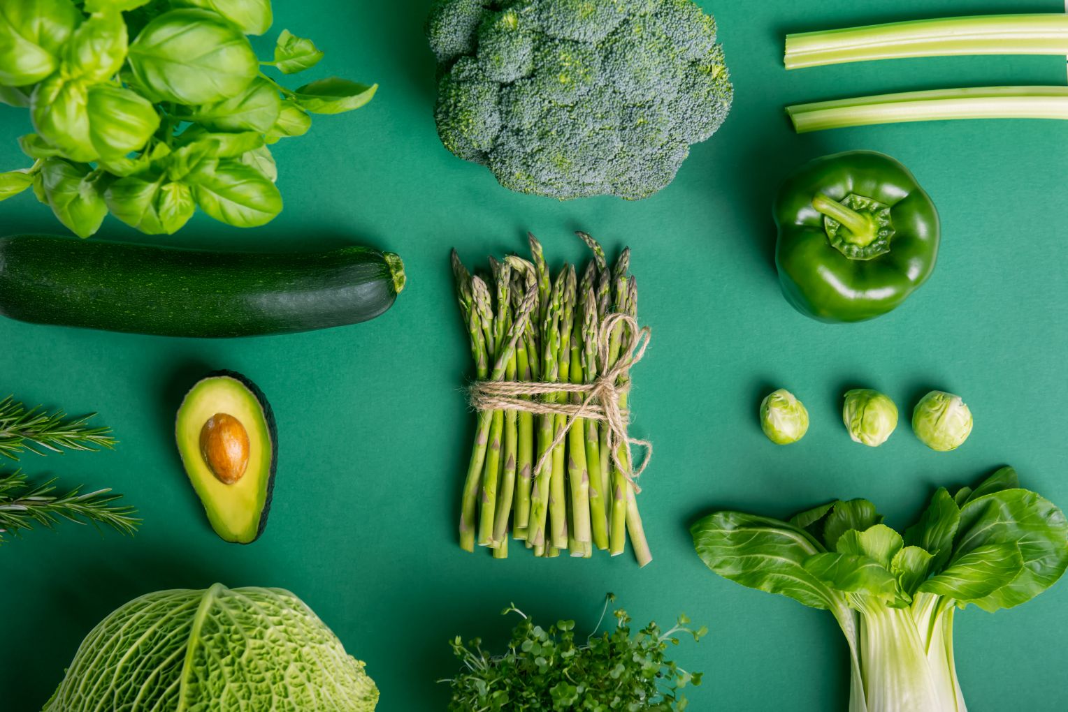 Verschiedene grüne Gemüsesorten wie Spargel, Brokkoli, Zucchini, Paprika, Avocado, Sellerie, Rosenkohl und Pak Choi sind kunstvoll auf einem grünen Hintergrund arrangiert – ideal für das Thema gesunde Ernährung.